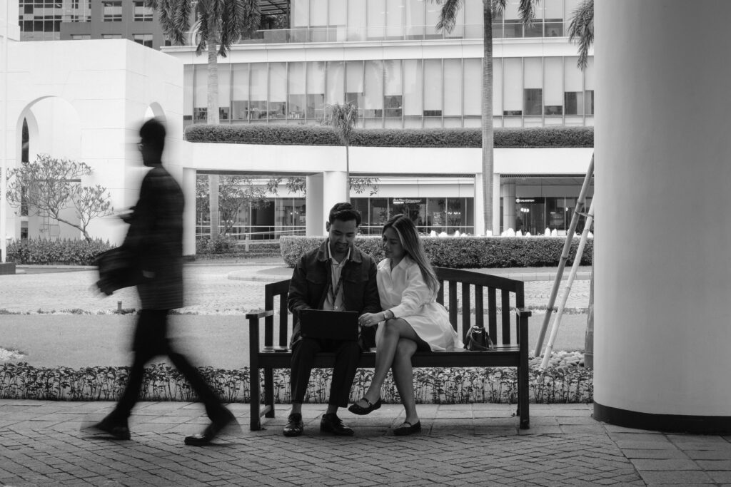 Black and white photo of a couple sitting on a bench in an urban plaza with palm trees, smiling while looking at a laptop together. A blurred figure walks past them.