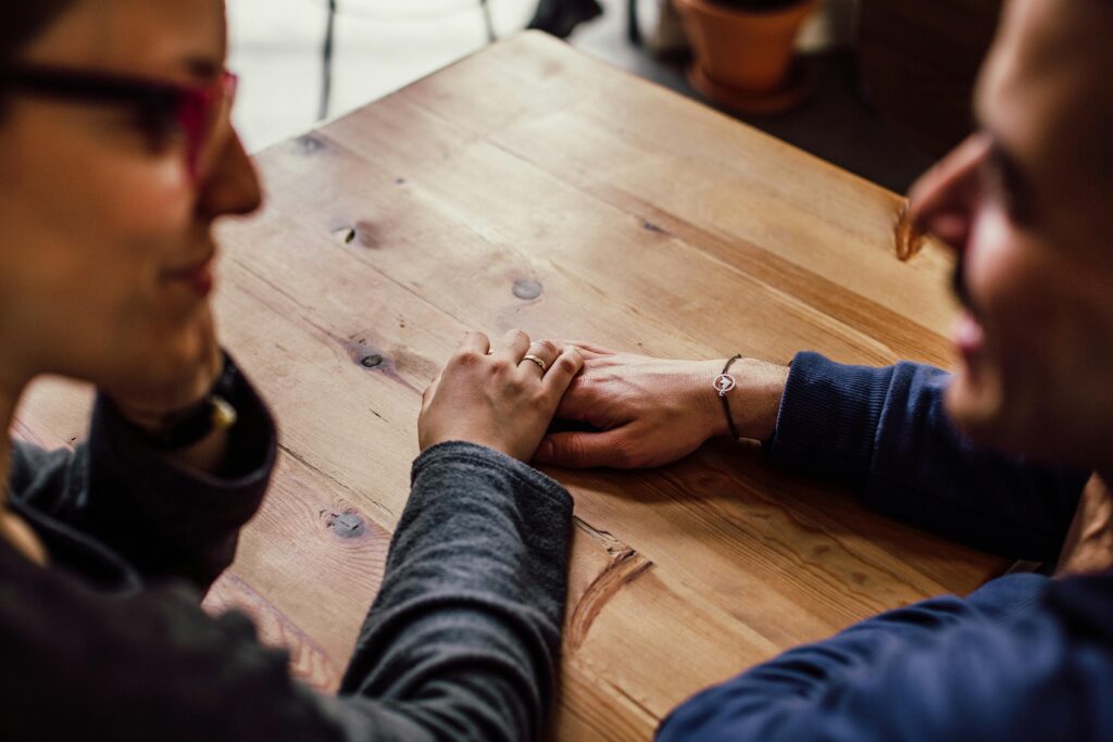 Two people hold hands across a wooden table, shot from a low angle with soft focus.