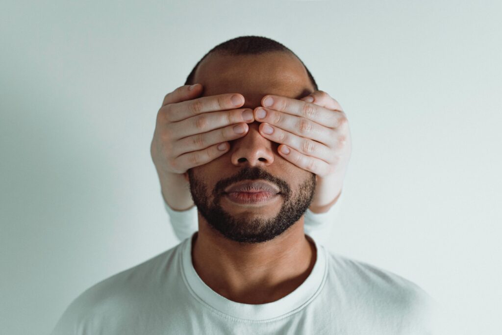 A bearded man in a t-shirt with someone else's hands covering both of his eyes against a plain light gray background.
