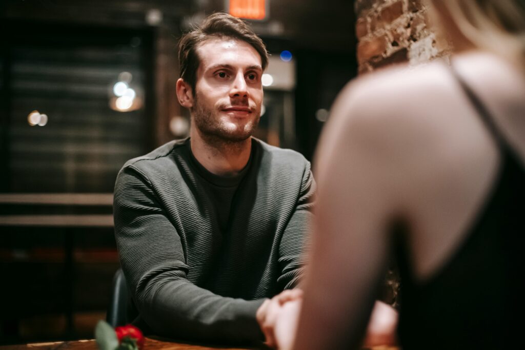 A man with brown hair and stubble in a dark sweater smiles softly at a woman seen from behind with blonde hair, in a dimly lit restaurant setting.