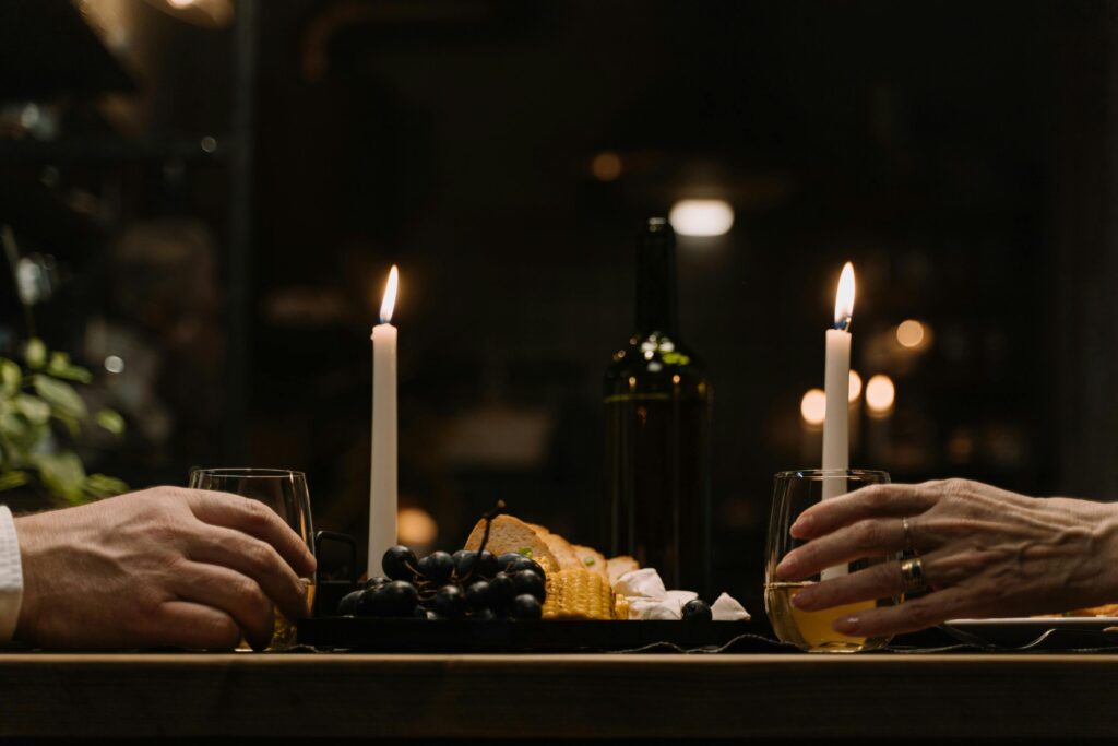 Two pairs of hands rest on either side of a candlelit dinner table with wine glasses, a wine bottle, cheese, and grapes between two lit taper candles.