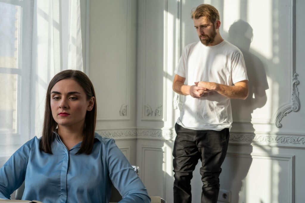 A woman in a blue button-up shirt sits looking away with a guarded expression while a bearded man in a white t-shirt stands behind her wringing his hands anxiously. Dramatic shadows from window light fall across the white ornate wall.
