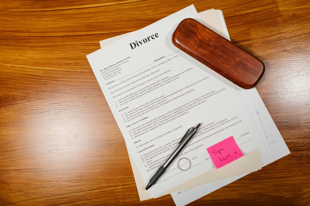 Divorce papers on a wooden desk with a silver pen, wooden pen case, and a pink sticky note reading "Sign here" with an arrow pointing to a signature line.