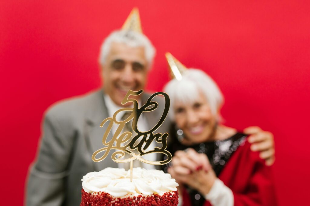 An elderly couple in gold party hats smiles behind a cake with a gold "50 Years" topper against a red background.