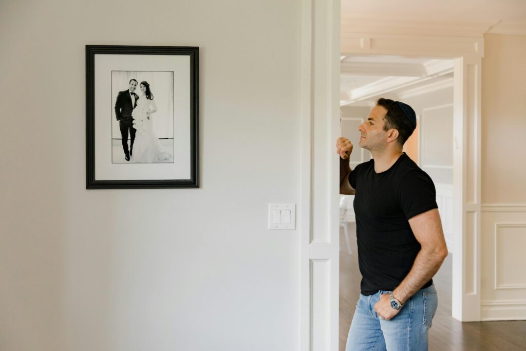 A man in a black t-shirt and jeans stands in a hallway gazing at a framed black and white wedding photo on the wall.