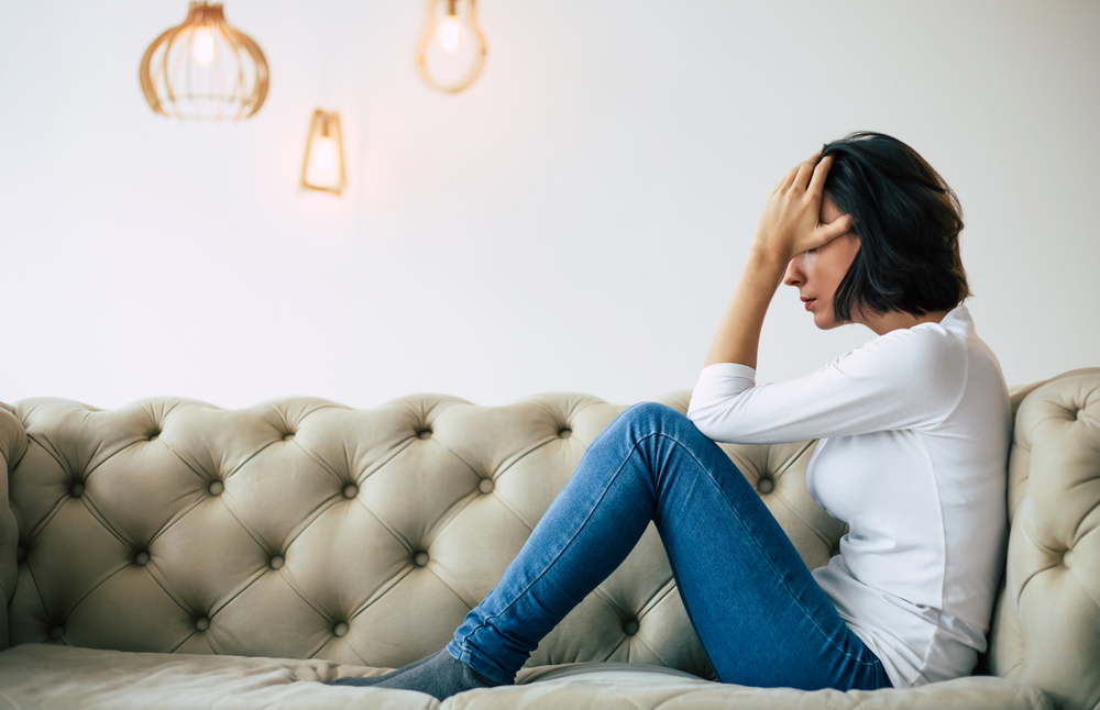 Chronic migraine. Close-up photo of a woman in casual clothes who is sitting on a sofa in her modern apartment, holding her head and grieving.