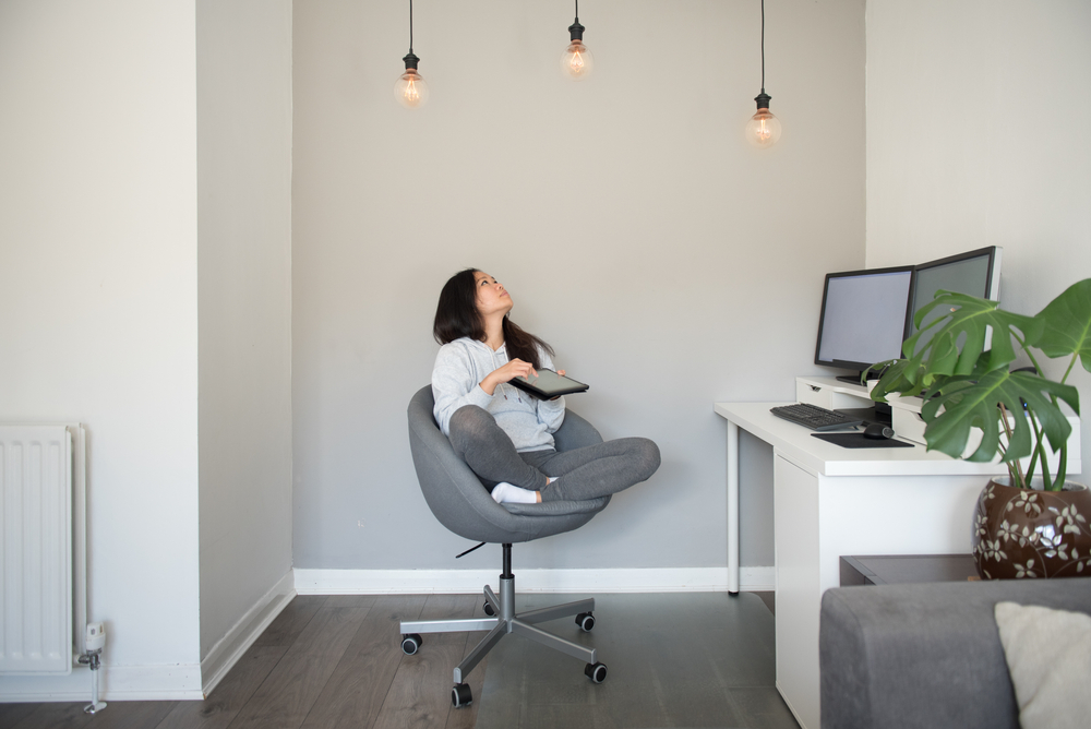 A young Asian woman adjusts the power of the ceiling LED lights from her digital tablet while working from home on her desktop computer sitting on an office chair in the living room of a house