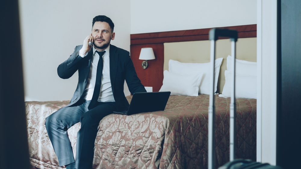Tilt up of smiling bearded businessman in suit working on laptop and talking at mobile phone while sitting on bed in hotel room. Business, travel and people concept