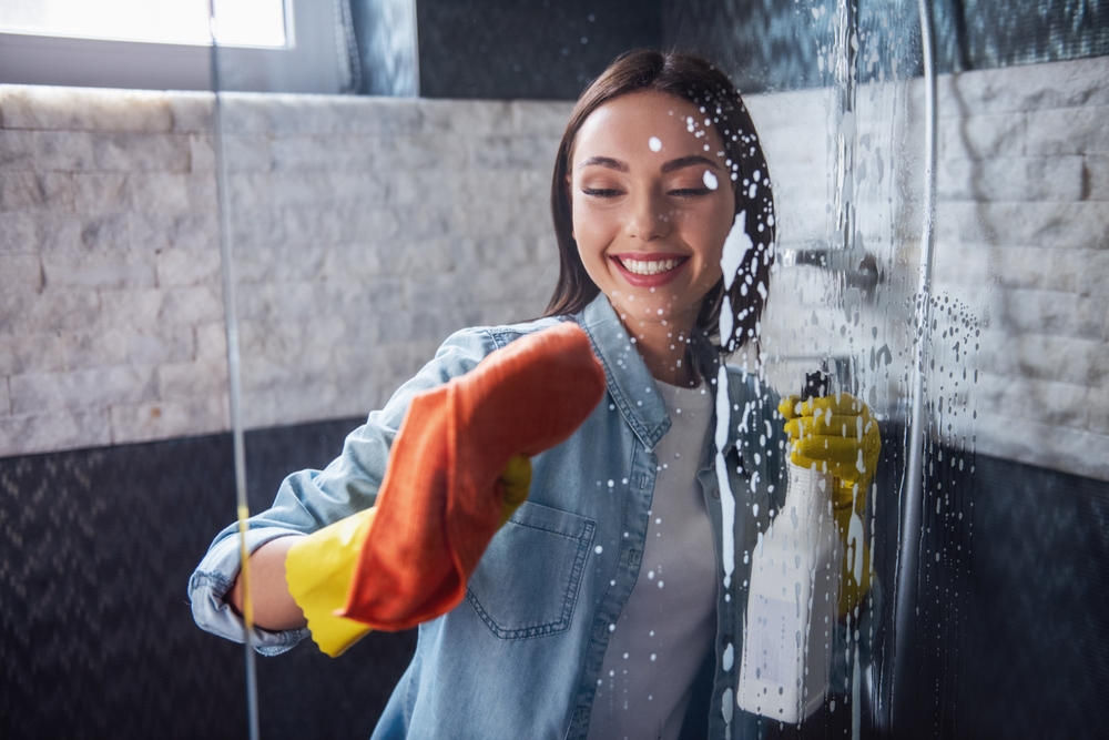 Beautiful young woman is using a detergent and a duster and smiling while cleaning a shower in bathroom