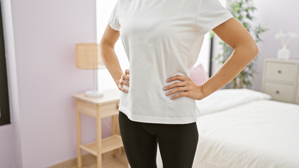 Cropped image of a young adult woman with hands on hips in a bedroom setting, portraying casual elegance and daily life.