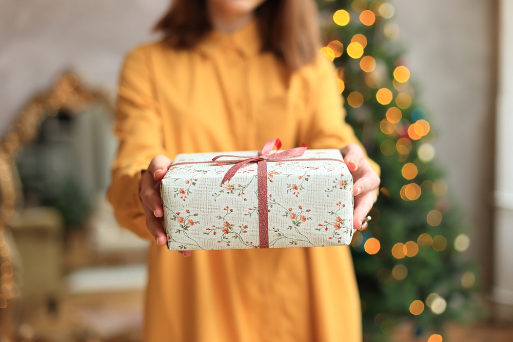 a young woman is joyfully opening a Christmas gift, wearing a bright yellow dress that radiates happiness. Girl holding gift wrapped box in hands on New Year's Eve