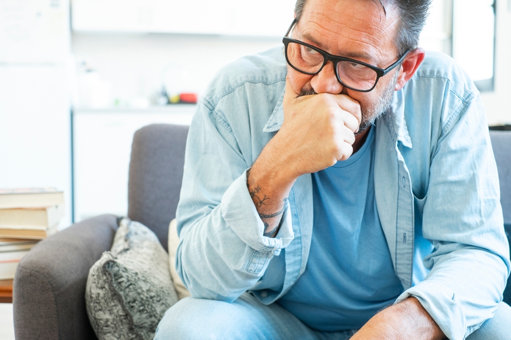 Mature individual reflecting on economic uncertainties, sitting in his living room with a somber expression, contemplating recent challenges with employment and relationship setbacks