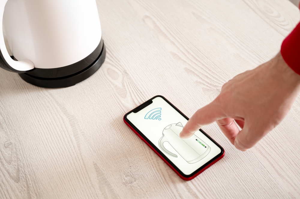 Hand using a smartphone to control a smart electric kettle, showcasing modern technology and convenience. Smart home