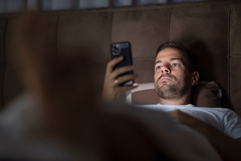 A man with serious facial expression and insomnia looking at cellphone and endlessly scrolling on cellphone in bedroom late at night.