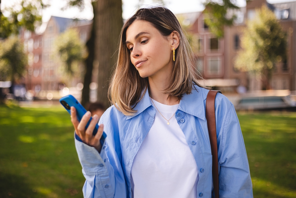 Mad and shock young beautiful woman using mobile phone standing in city in park. Unhappy woman hold mobile phone. Disappointed sad upset lady horrified impressed news. WTF. Oh no, roll eyes.