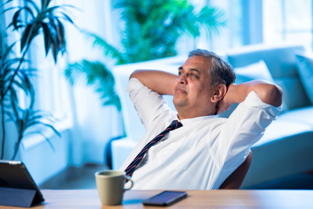 Indian businessman relaxing on chair at office desk in modern workplace, Asian corporate executive leaning back with calm expression taking a short break during workday, symbol of stress relief