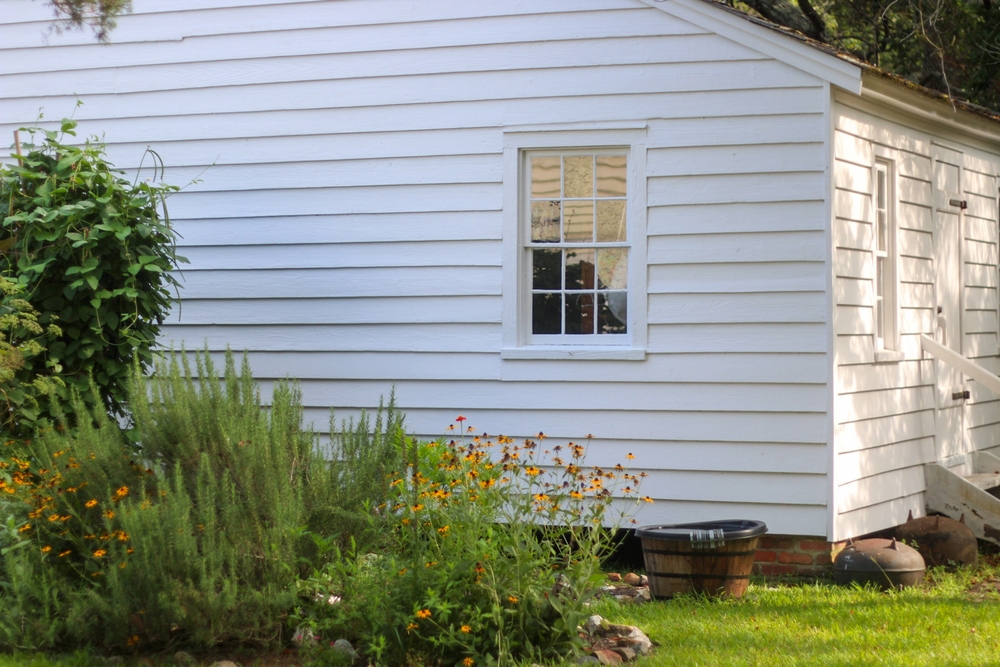Old building with lap siding wavy glass window and a small garden of black eyed Susans. 