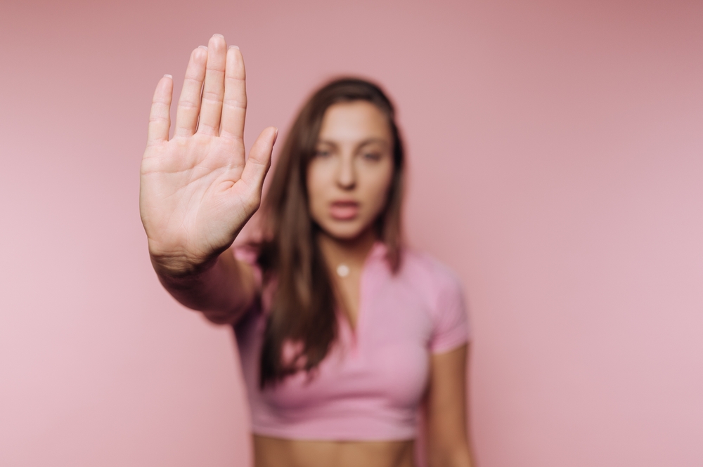 Woman in pink shirt holding hand up in stop gesture on pink background.