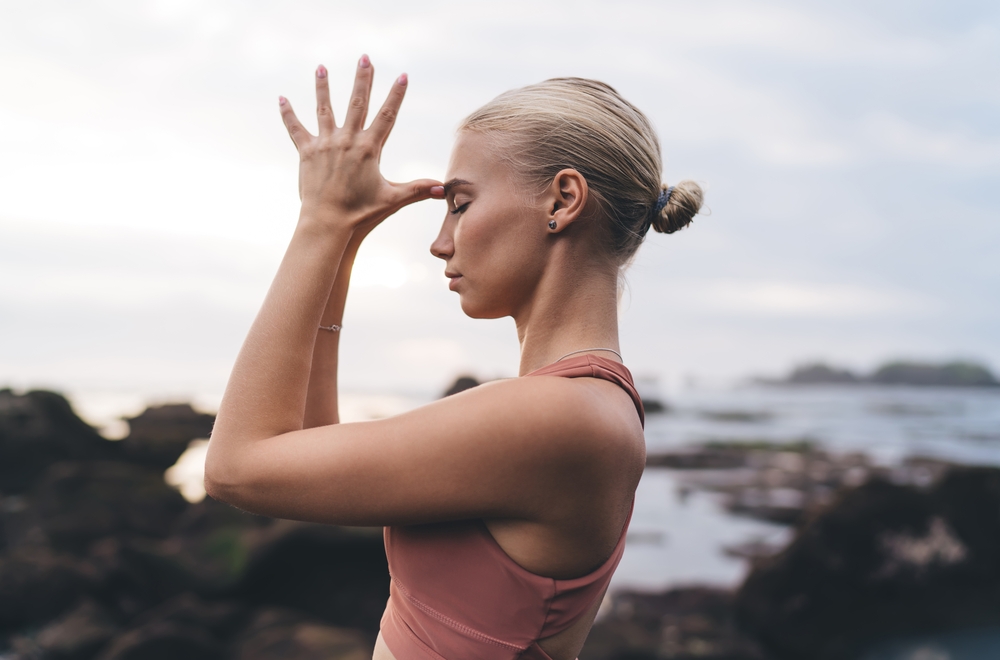 Profile view of young woman in yoga wear touching forehead with fingers, eyes closed, symbolizing intuitive vision, inner focus and wellness as resistance to tech saturation.