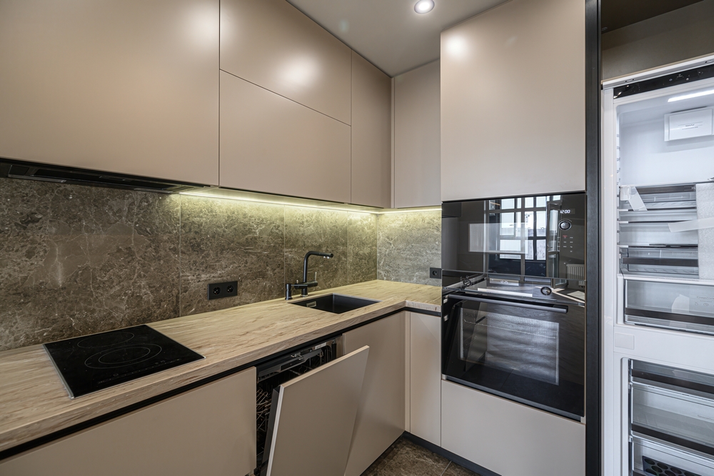 Modern kitchen. Beige cabinets, marble backsplash, black sink faucet, oven, open dishwasher, and fridge. Under-cabinet lighting