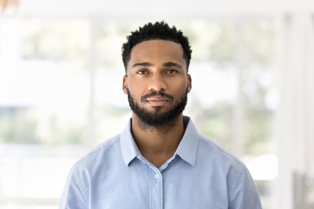 Head shot home portrait of young African man wear blue casual shirt standing alone on sunny blurred background. Millennial generation person profile picture. Tenant or homeowner portrait, independence