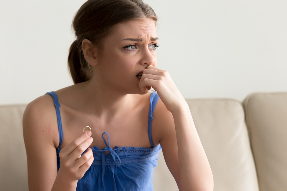 Doubtful young woman holding wedding ring in hand, feeling unsure hesitating about engagement proposal, sad wife thinking about breaking up or getting divorced concept, unhappy marriage