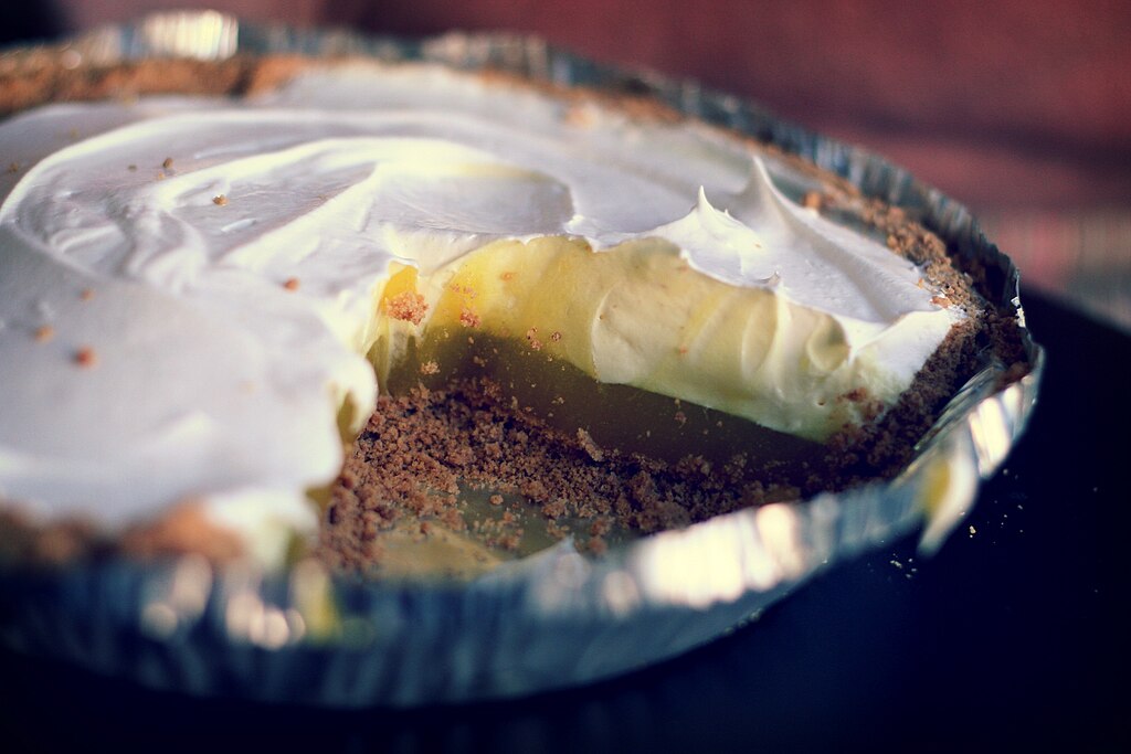 A pie with graham cracker crust and yellow pudding filling, topped with white whipped cream, with one slice missing.