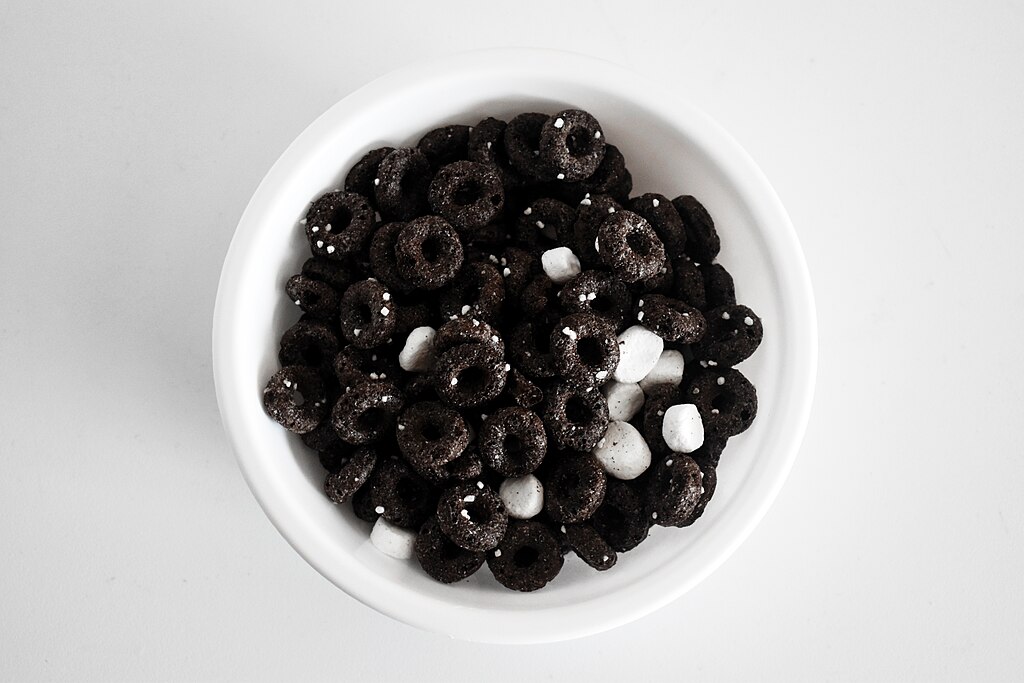 A white bowl of chocolate ring-shaped cereal with small white marshmallows, viewed from above on a white background.