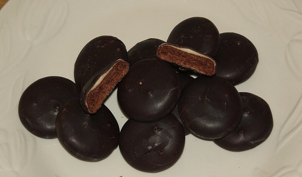 Dark chocolate-covered round cookies on a white plate, with two broken open to show the chocolate cookie interior and thin cream filling.