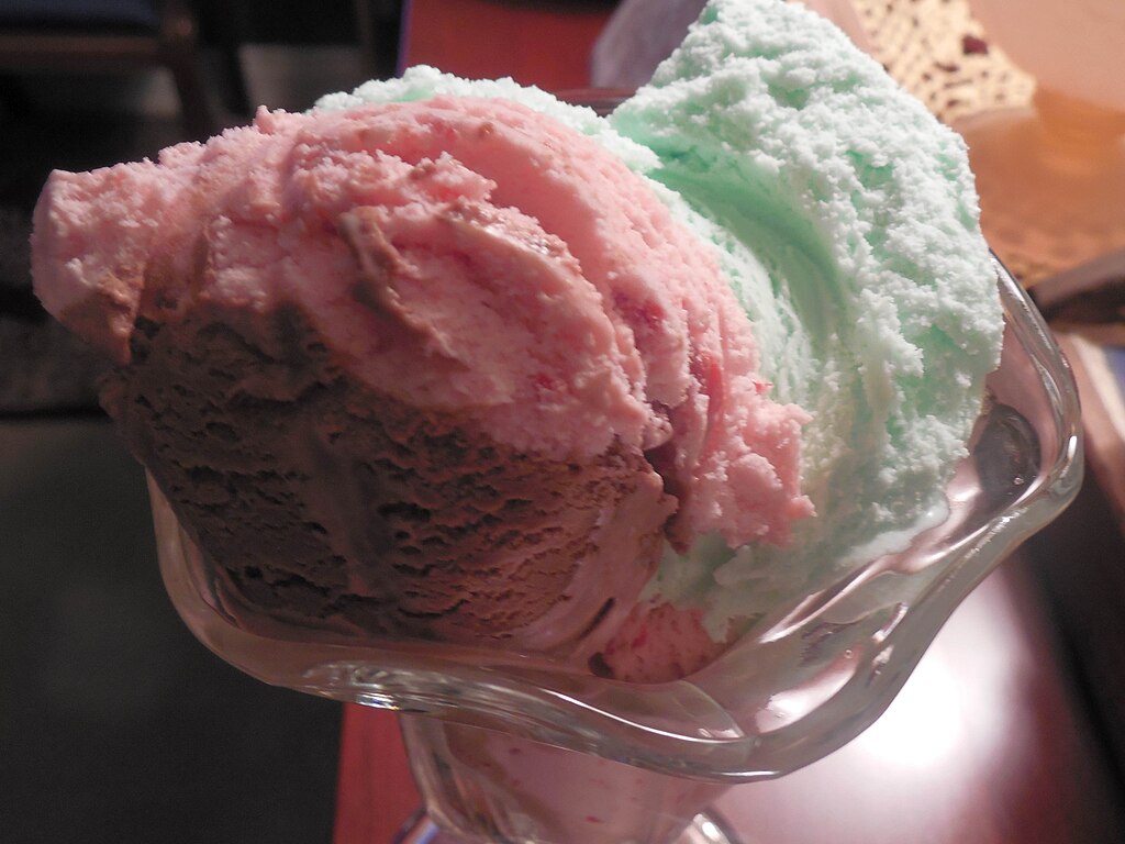 Scoops of pink, green and brown spumoni ice cream in a glass dessert dish.