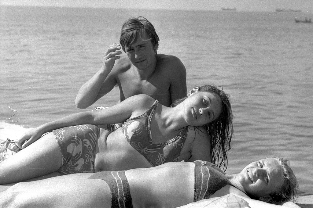 A black-and-white photo of three young people relaxing at a beach, with a woman in a patterned high-cut bikini leaning against a man in swim briefs while another woman lies in the foreground.