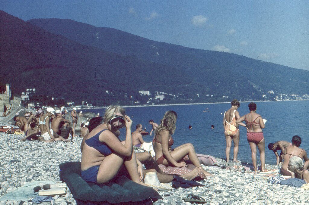 A color photo of a crowded pebbly beach with forested mountains rising in the background, showing dozens of people in swimsuits sitting, standing, and sunbathing along the Black Sea shore.