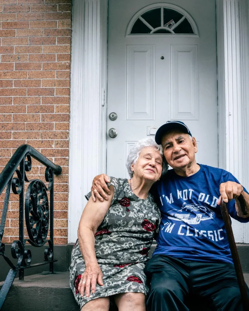 An elderly couple sits on brick front porch steps. The woman leans contentedly against the man, who wears a t-shirt reading "I'm not old, I'm classic."