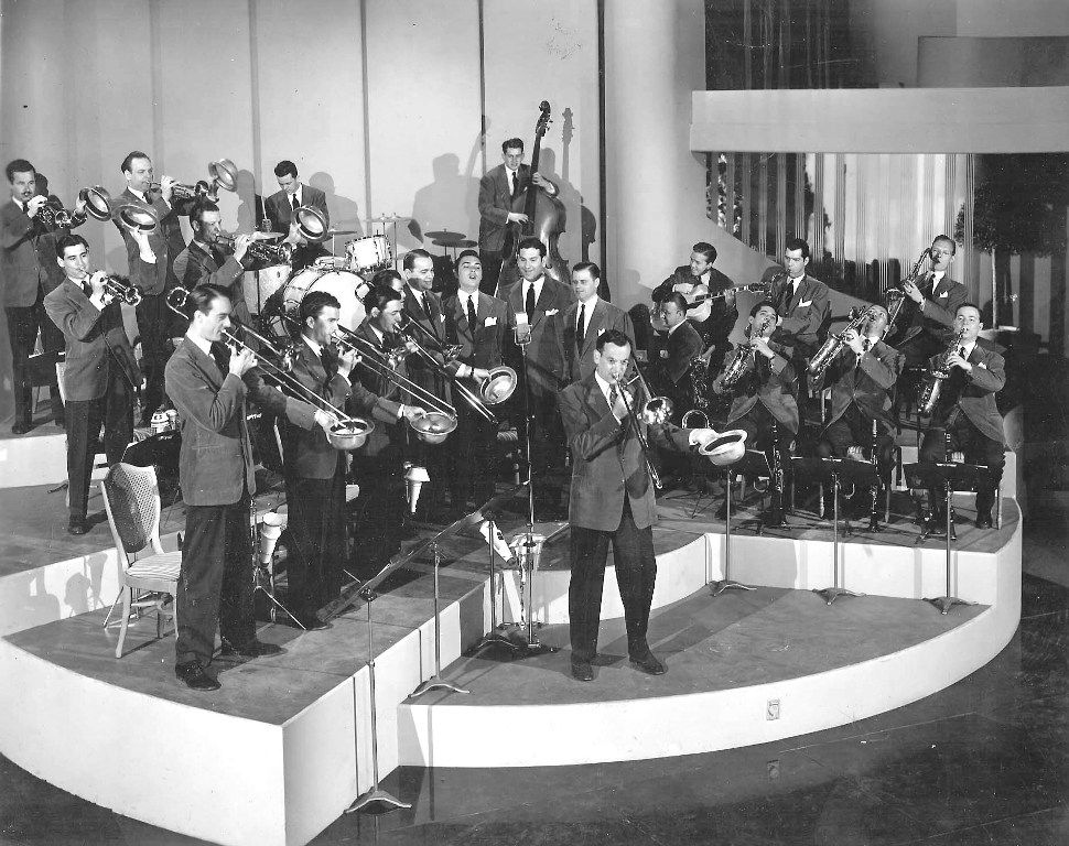 Black and white photograph of the Glenn Miller Orchestra performing on a curved, tiered stage, with the bandleader standing center front at the microphone while rows of musicians in suits play brass instruments behind him and the rhythm section visible in the back.