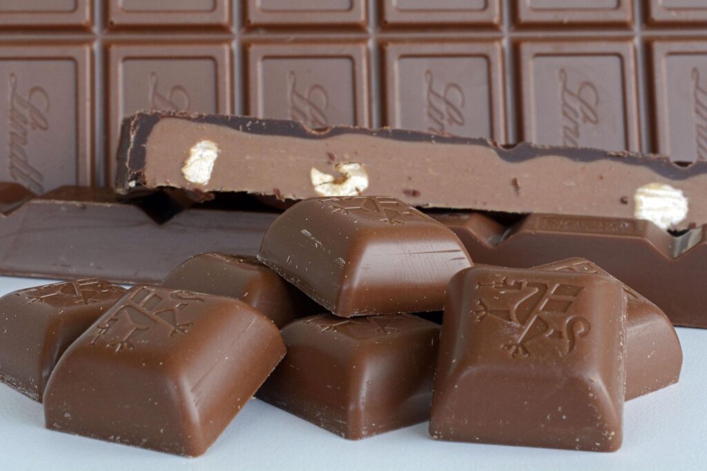 Milk chocolate bar with broken squares in foreground, showing embossed brand logo on each piece.