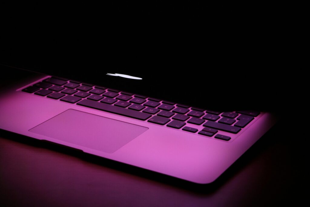 Laptop keyboard and trackpad illuminated by pink light from the screen in a dark room.