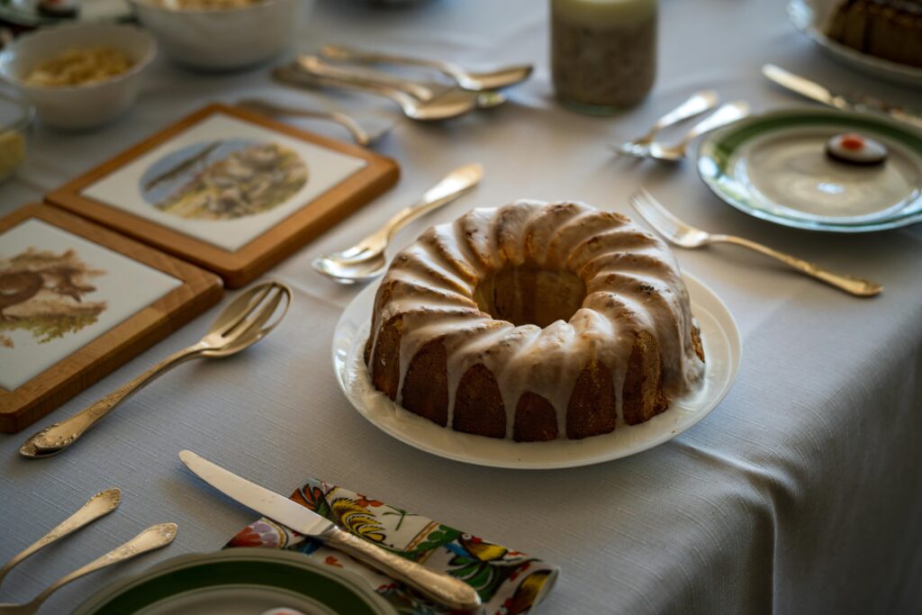 A glazed bundt cake with white icing dripping down the sides, displayed on a set dining table with utensils and serving dishes.