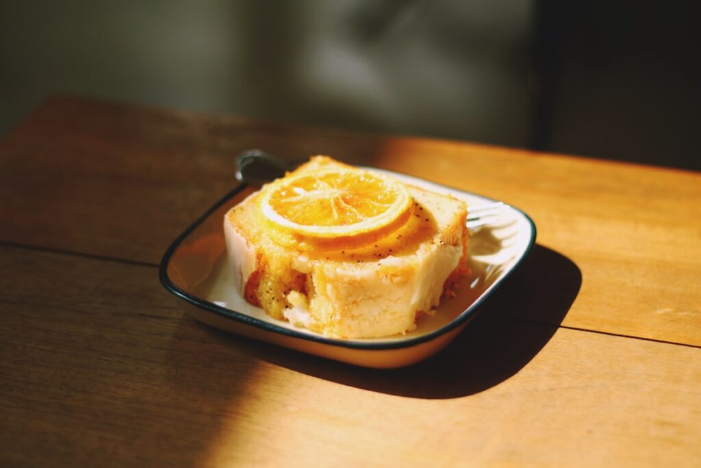A square slice of golden yellow cake topped with candied orange slices in a small enamel dish on a wooden table.