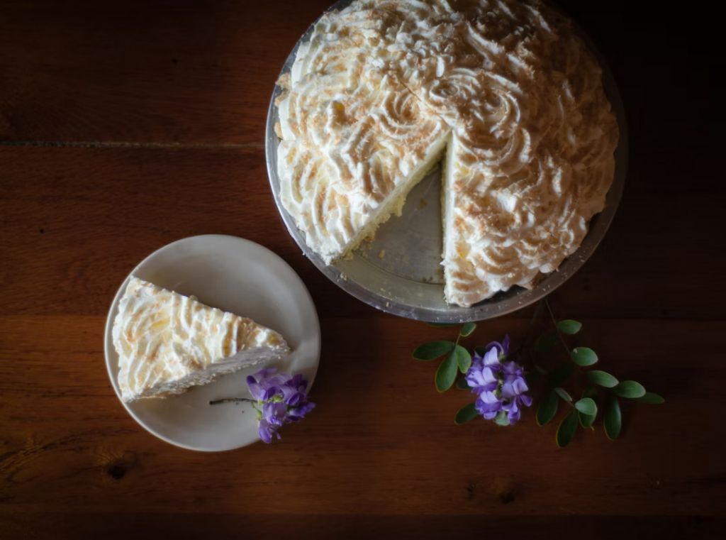 A whole coconut cream pie  on a cake stand, one slice served on a small plate beside purple flowers on a wooden table.