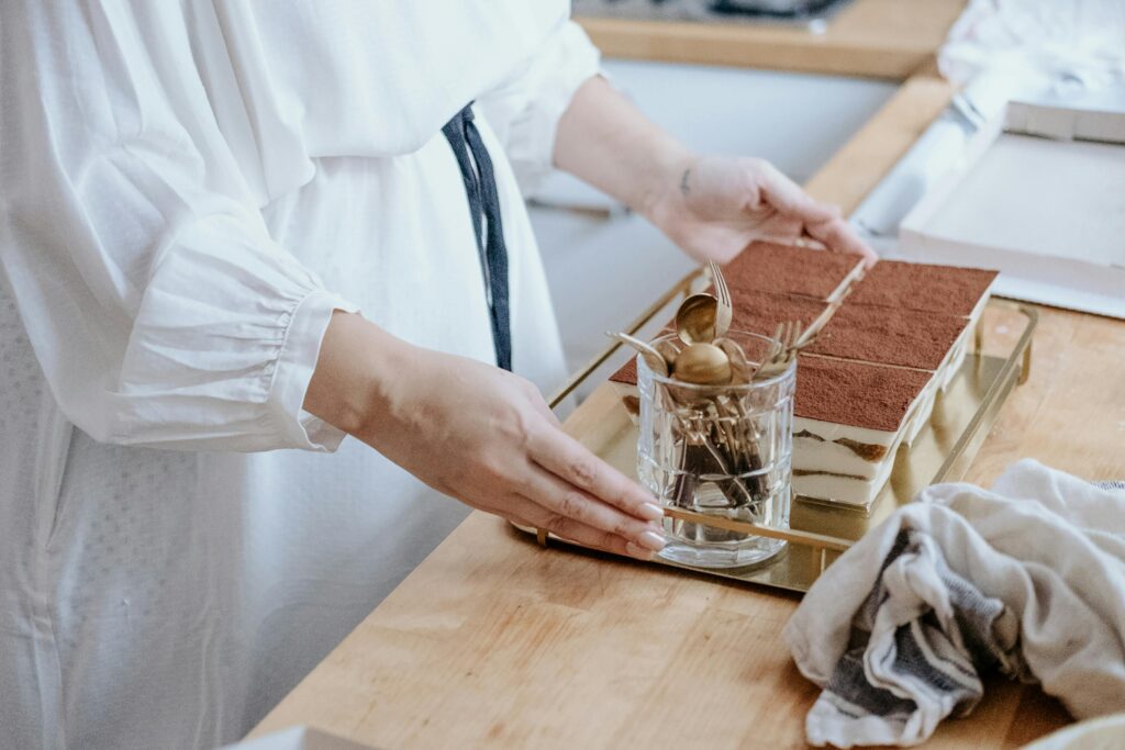 A person in a white blouse arranging a rectangular tiramisu dusted with cocoa powder on a wooden kitchen counter.