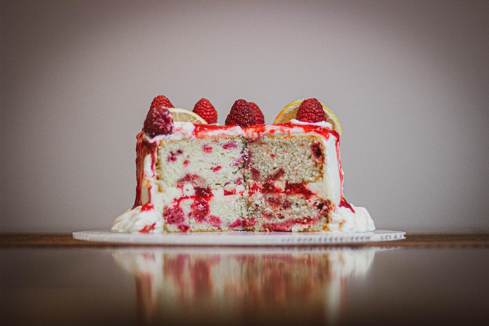 A sliced layer cake showing red Jell-O streaks throughout white cake, topped with white frosting, fresh raspberries and lemon slices.