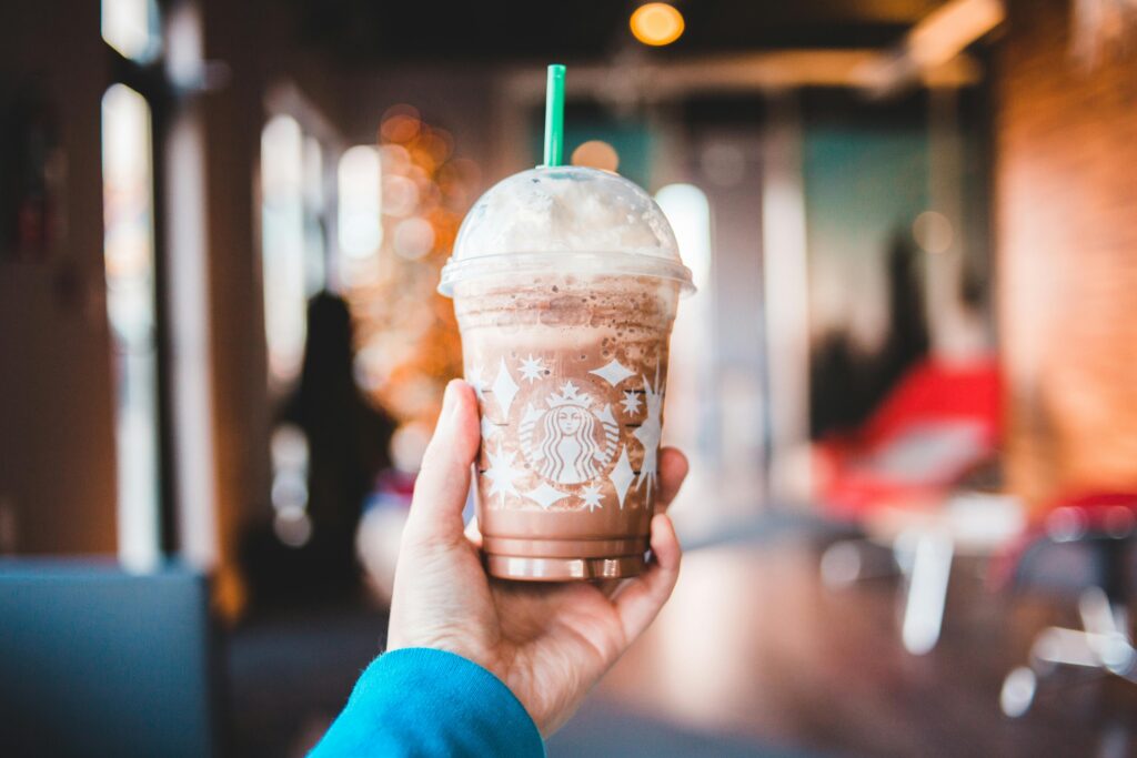 A hand holding a Starbucks Frappuccino topped with whipped cream, with a blurred café background and warm bokeh lights.