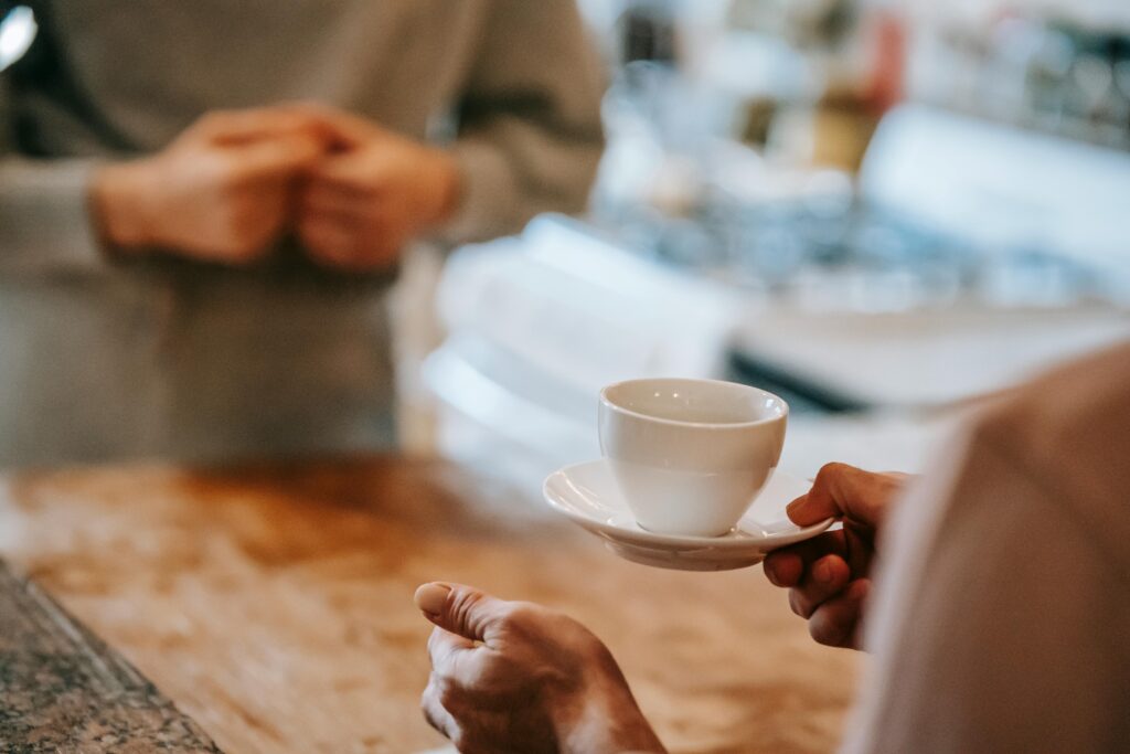 Hand holding a white coffee cup and saucer at a cafe counter with a blurred figure in the background.