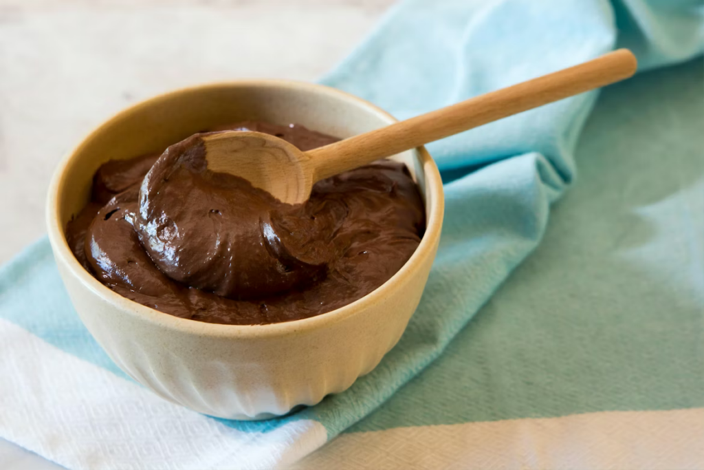 A yellow ceramic bowl filled with dark chocolate mousse and a wooden spoon, resting on a blue cloth napkin.
