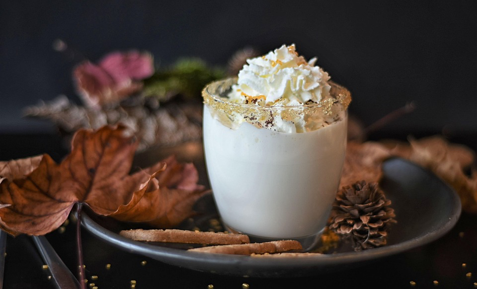 A mug of white chocolate mousse topped with whipped cream, surrounded by autumn leaves and cinnamon sticks on a dark background.