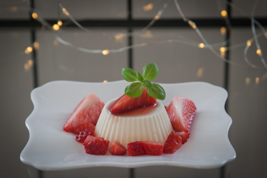 A molded white panna cotta surrounded by fresh strawberry slices and topped with basil on a decorative plate, with twinkling lights behind it.