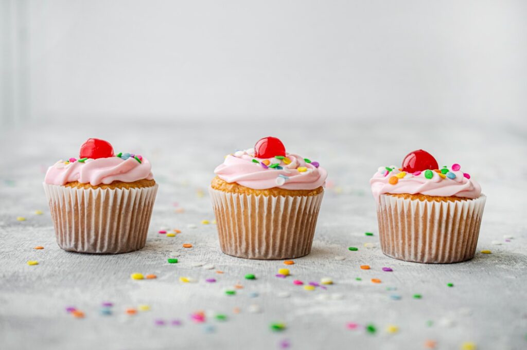 Three vanilla cupcakes with pink frosting, colorful sprinkles and maraschino cherries on a light gray surface.