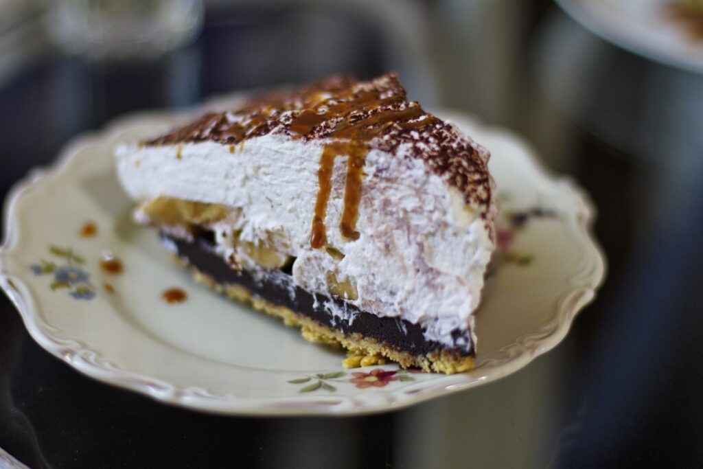 A layered dessert slice on a floral china plate, showing chocolate pudding, whipped cream and a cookie crust with caramel drizzle.