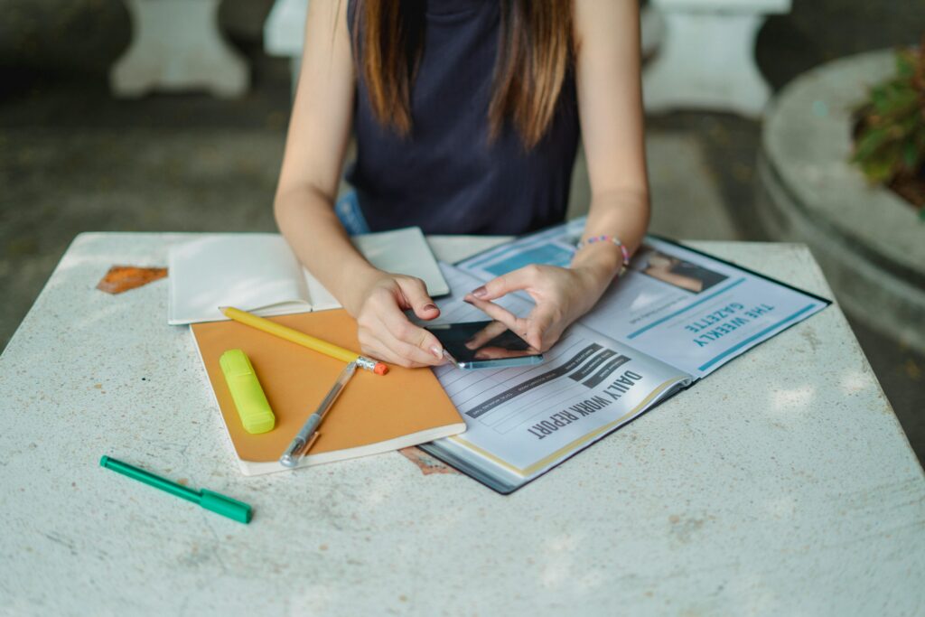 A teenage girl uses her smartphone while doing homework at a table with notebooks, pens, and open textbooks.