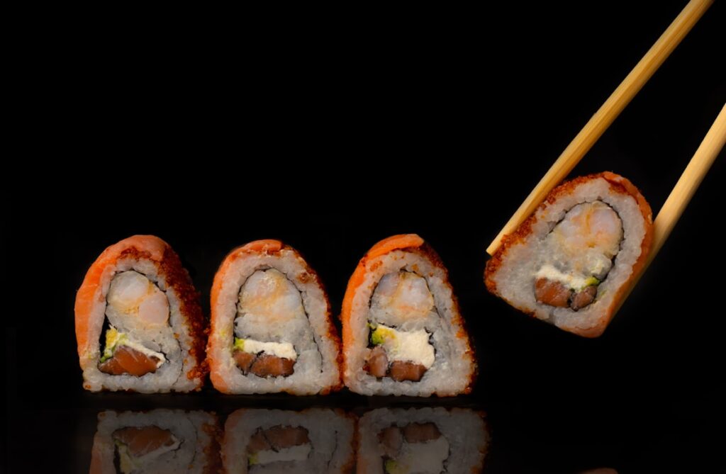 Four salmon sushi rolls with shrimp and avocado on a reflective black surface, one piece held by wooden chopsticks.